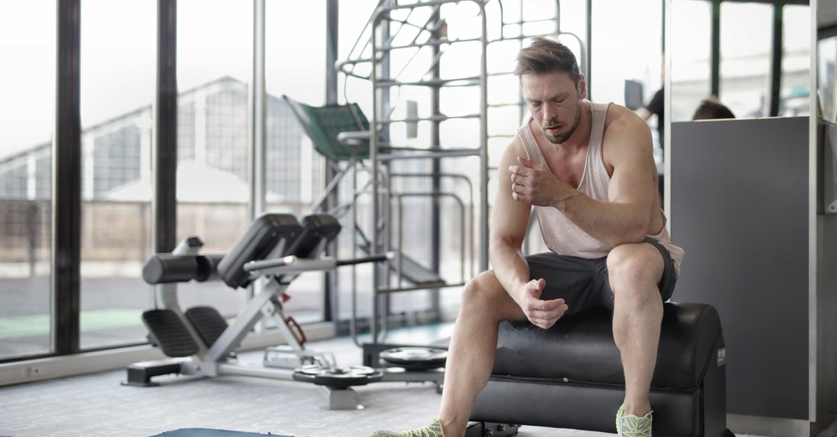 Tired young muscular male athlete in casual sportswear and sneakers sitting on bench and resting after intense weightlifting workout in modern fitness club